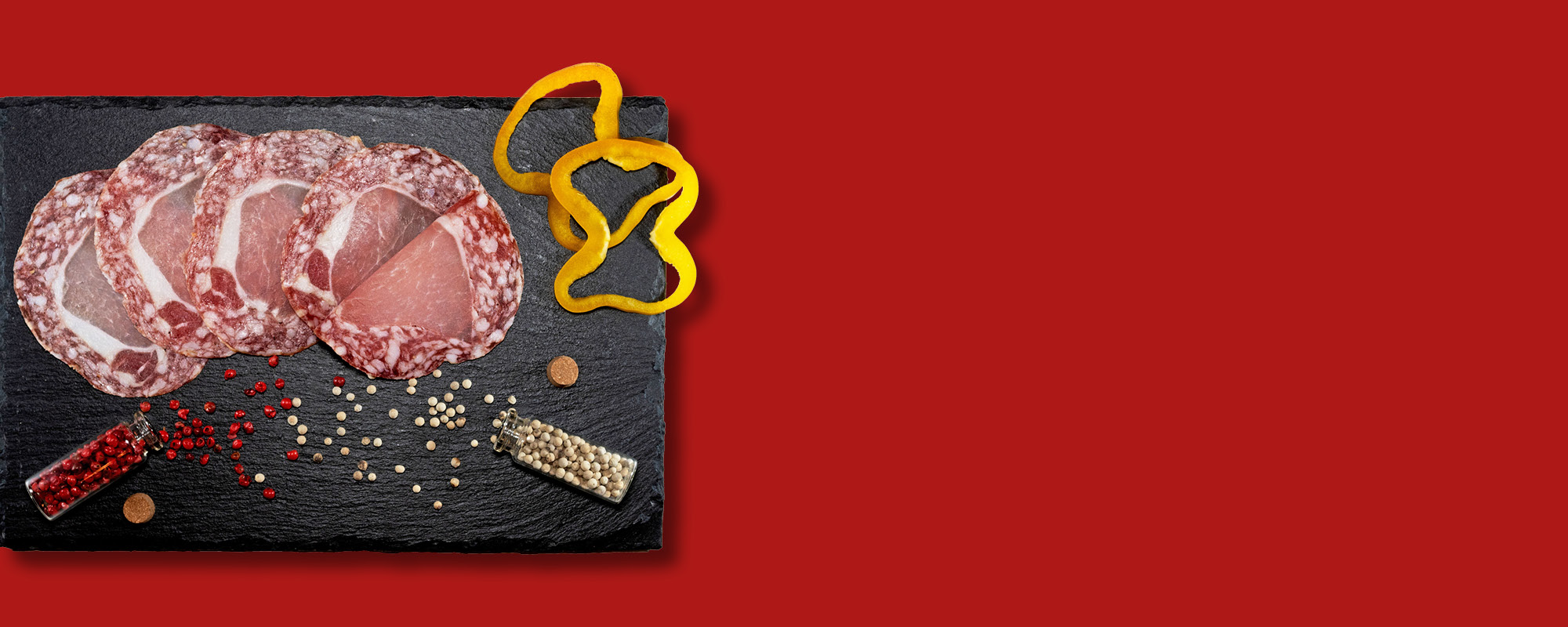 Fette di salame su una pietra ardesia scura, circondate da grani di pepe rosa e bianco, fette di peperone giallo e una boccetta di vetro, il tutto su sfondo rosso.