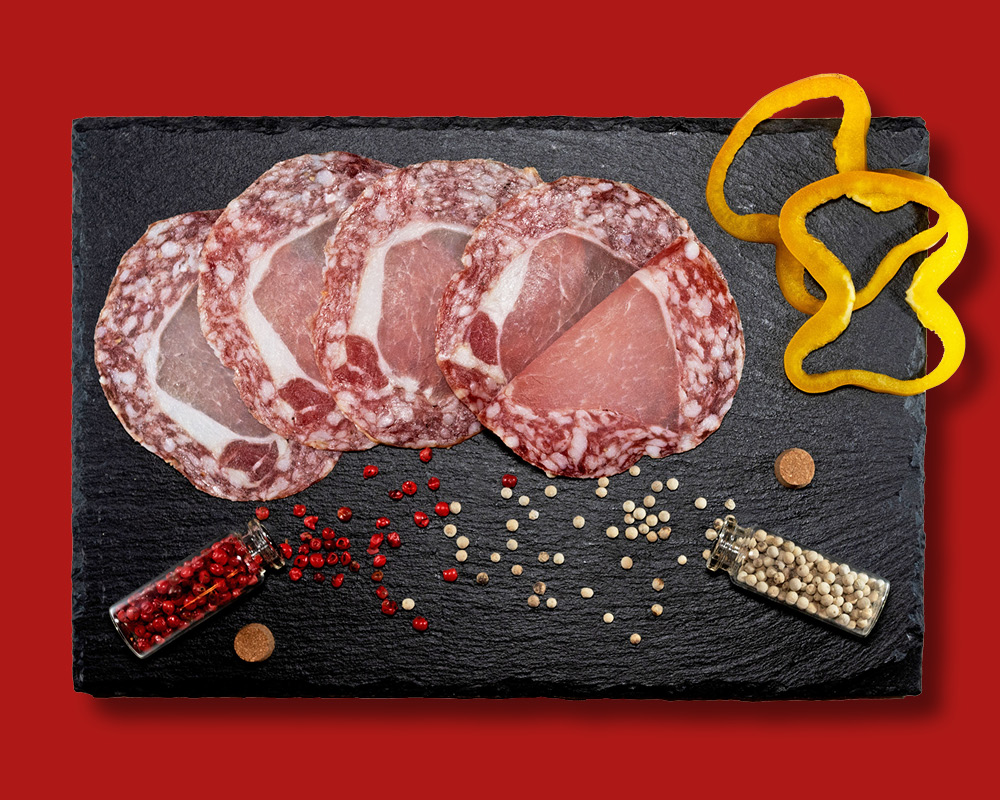 Salumi affettati su una pietra ardesia, accompagnati da fettine di peperoni gialli, spezie rosse sparse e un barattolino di pepe rosa