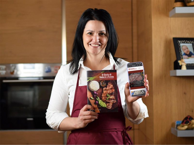 Una donna dai capelli neri sorridente indossa un grembiule rosso e tiene nella mano destra un libro di cucina intitolato "Il Gusto di Sedersi a Tavola". Nella sinistra ha uno smartphone che mostra la sua app con la scritta "LE NOSTRE RICETTE". Lo sfondo è una cucina moderna con un forno e delle foto di famiglia visibili su delle mensole.