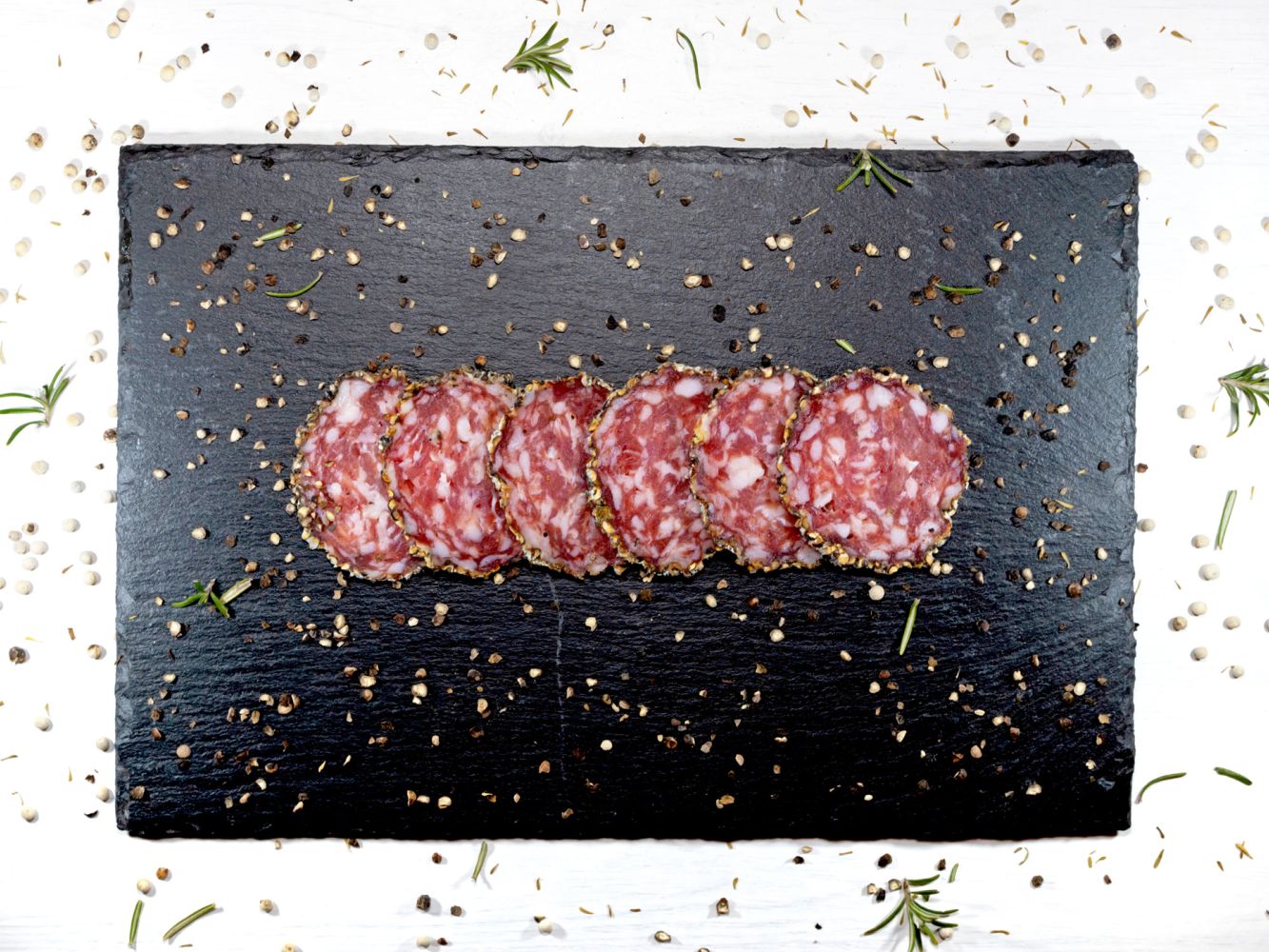 Fette di salame con bordo speziato disposte su un tagliere nero, circondate da grani di pepe e rametti di rosmarino.